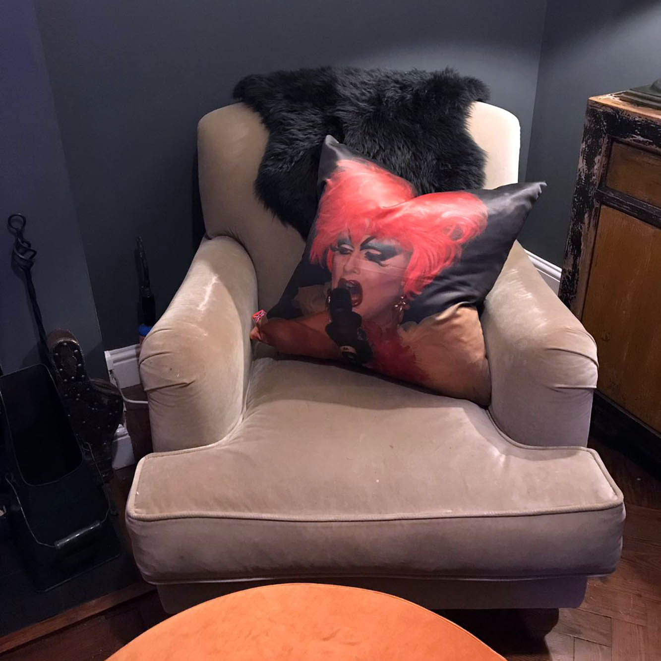 Drag queen cushion by Claire Swindale resting on taupe velvet armchair with black sheepskin throw