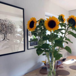 original painting of a willow tree, framed, on wall, next to vase of large sunflowers